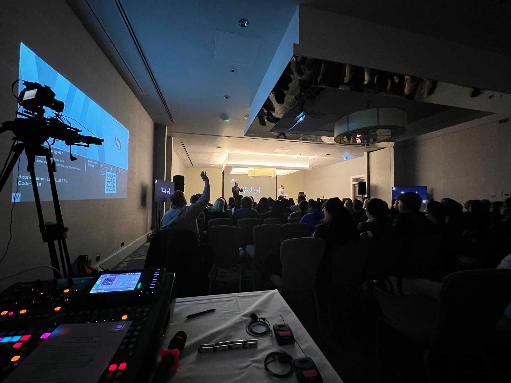 Conference presentation setup with AV equipment, including sound mixer and camera, capturing audience engagement in a dimly lit room.