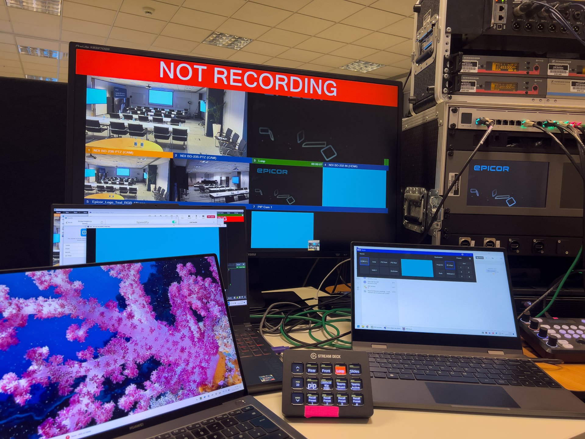 AV control room setup with multiple screens displaying a conference room feed, equipment racks, and sound mixing desk.