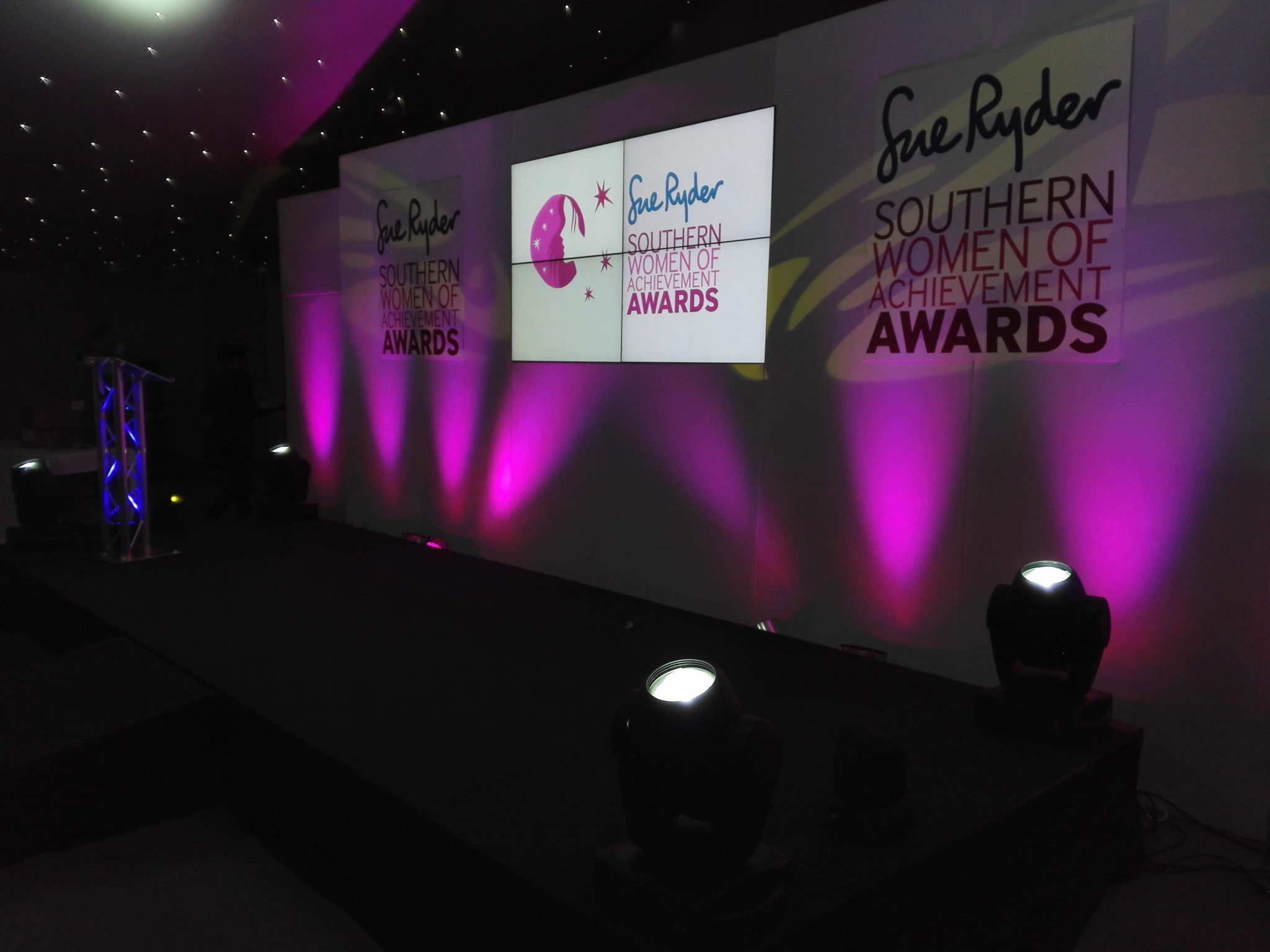 Stage setup with pink uplighting for the Southern Women of Achievement Awards, featuring podium, event backdrop, and logo display.