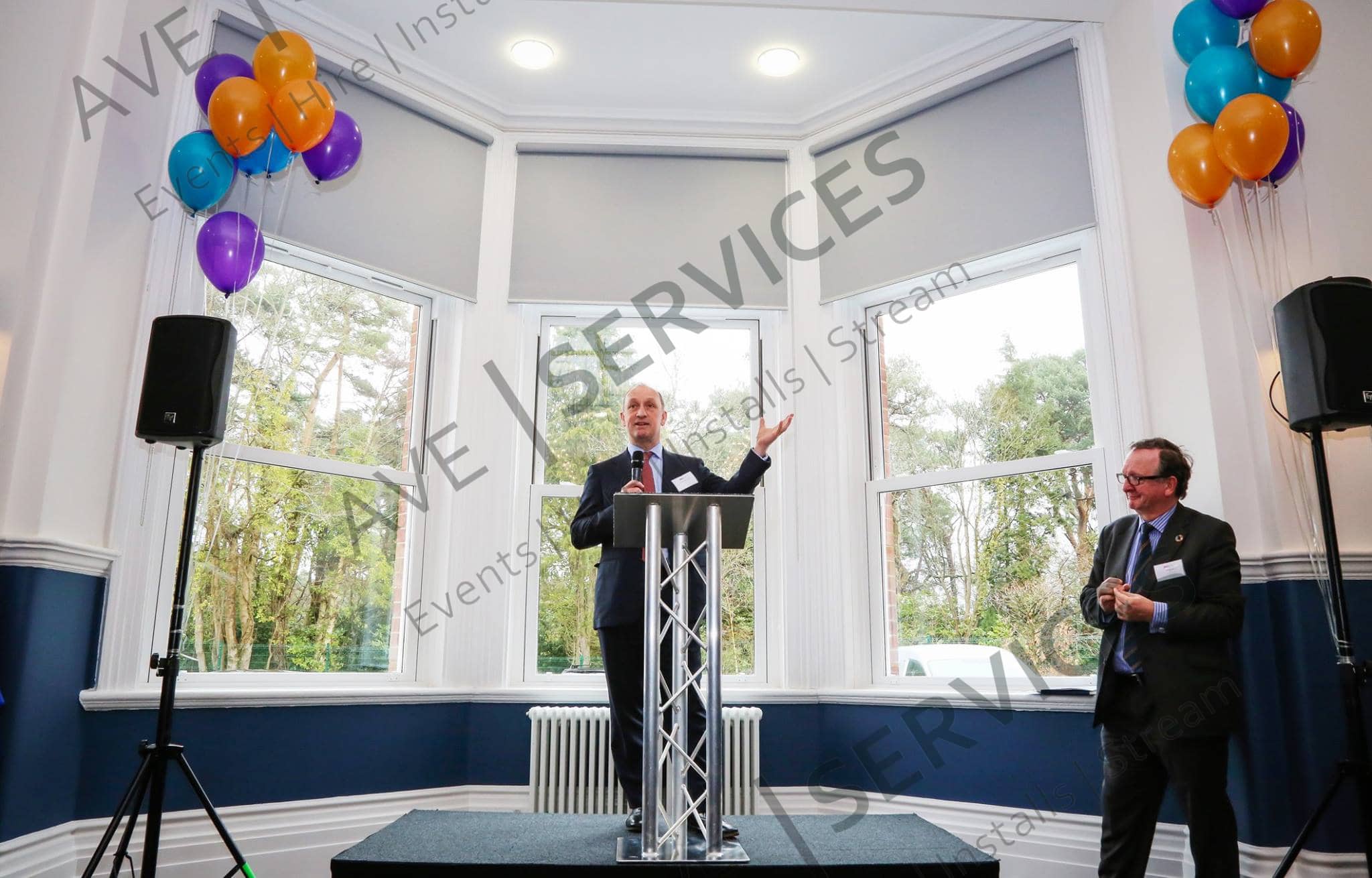Speaker presenting at a corporate event with balloon decorations and AV sound setup, featuring staging and indoor lighting.