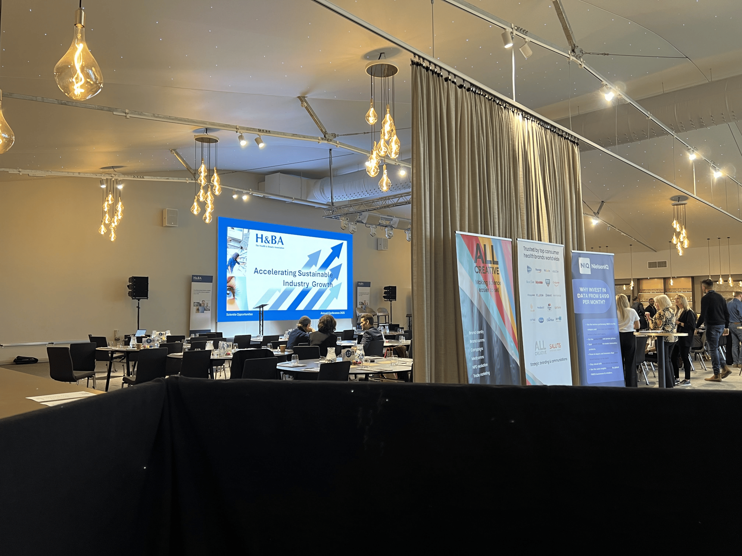 Corporate conference setup with staging, screen projection, and lighting. Attendees gather around tables with promotional banners visible.