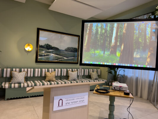 Conference room featuring a large projector screen, podium with Shalem College logo, and seating area with striped cushions.
